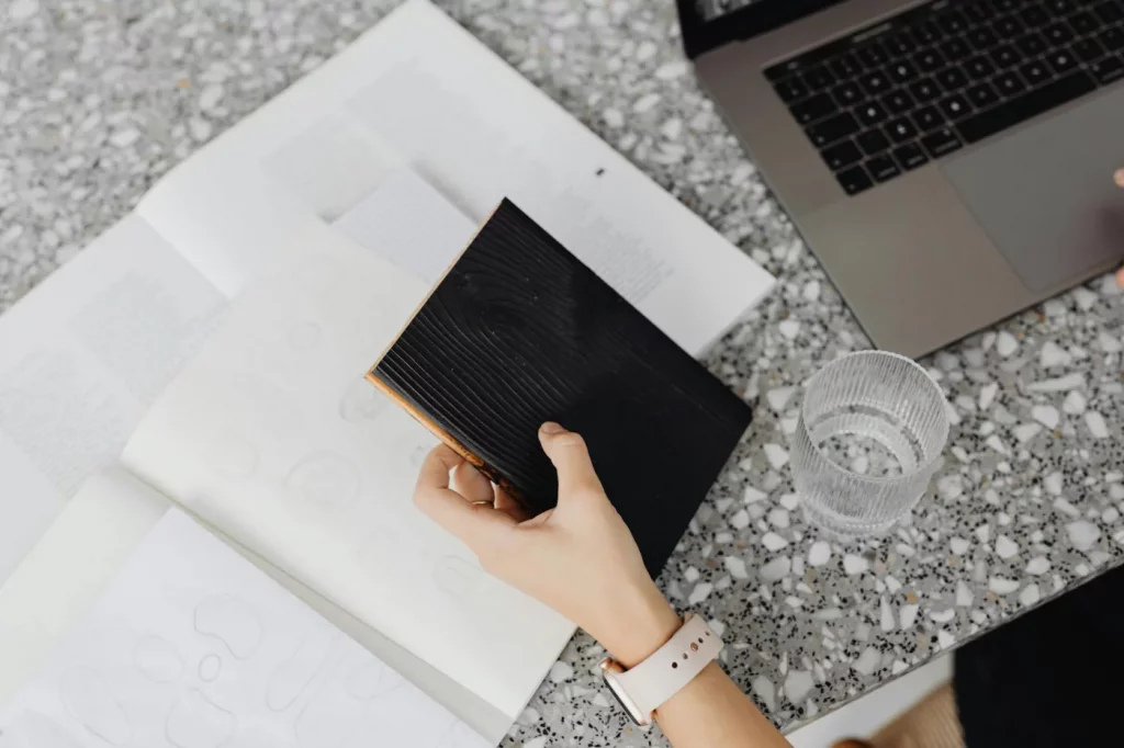 Hand holding black notebook by laptop and water, planning tasks with getting things done gtd guide pdf