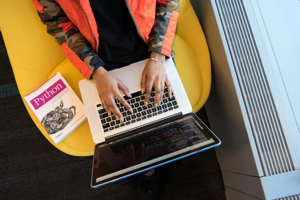 Woman coding on a laptop with a Python book nearby, illustrating how to learn python faster in 30 days