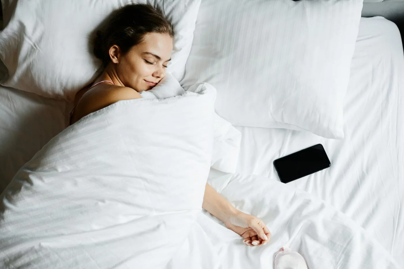 How to Build a Bedtime Routine for Adults That Helps You Fall Asleep Faster Bedtime routine for adults: a woman sleeping peacefully in white sheets with a smartphone beside her
