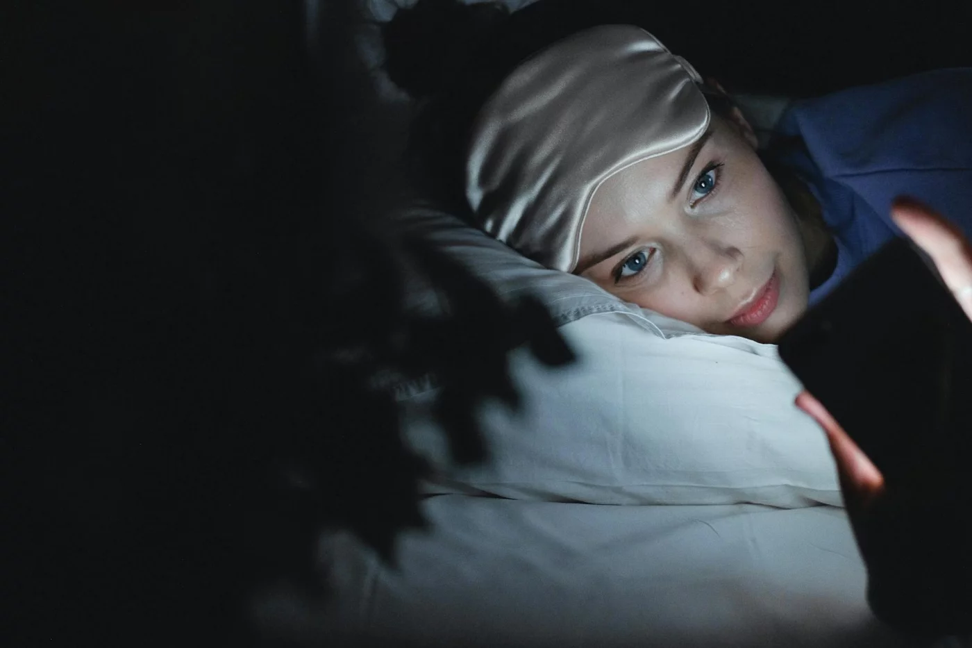 Blue Light and Sleep: How to Protect Your Brain at Night Woman in bed wearing a sleep mask uses her phone at night, showing how does blue light affect sleep quality