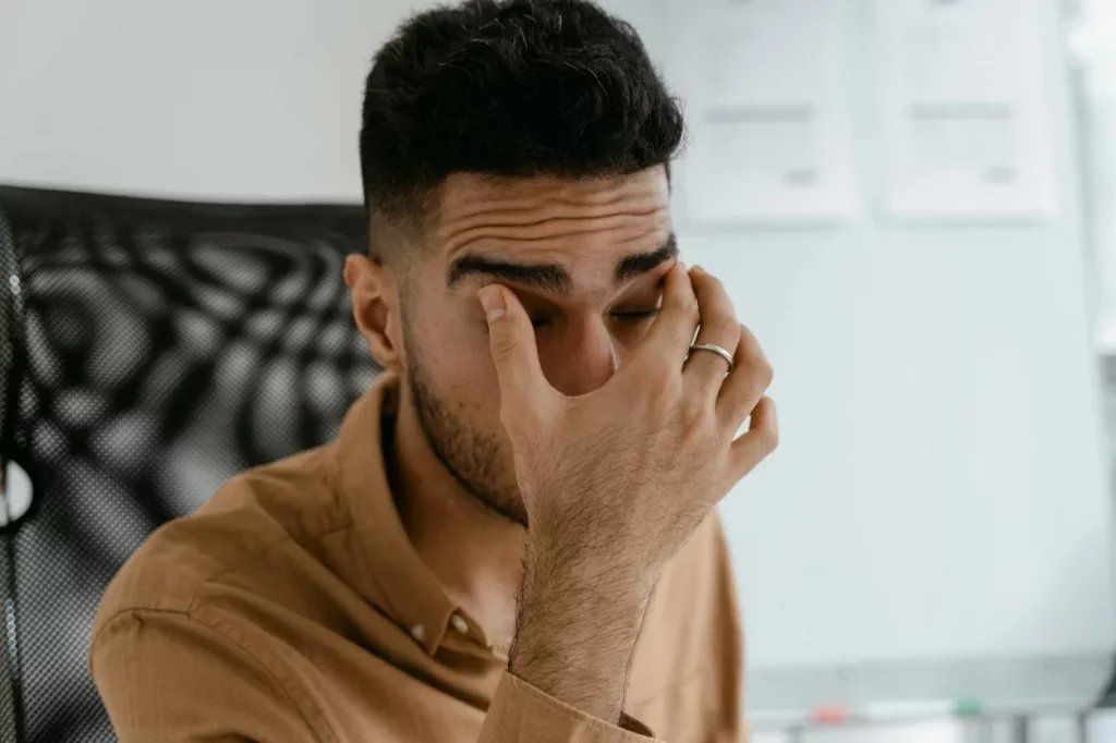 Office worker rubbing his eyes at a desk, showing physical symptoms of burnout and stress-related fatigue